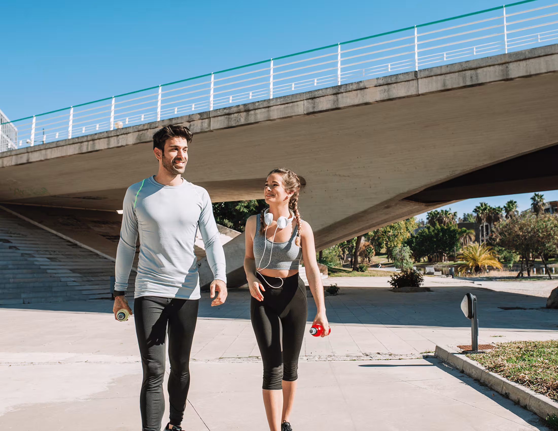 A young man and woman in sports clothing walking side by side with a bridge in the background.